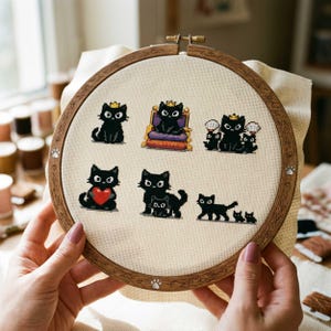 Peut inclure: Un cercle à broder au point de croix avec plusieurs motifs de chats noirs. Les chats sont représentés dans différentes poses, certains portant des couronnes ou tenant des cœurs. La broderie est fixée dans un cercle en bois.