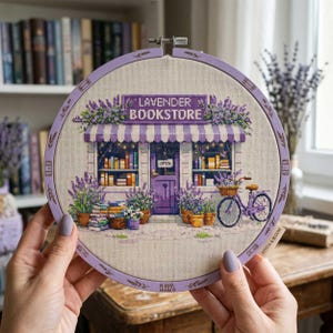 Könnte beinhalten: Ein Kreuzstich-Stickrahmen mit einer Lavendel-Buchladen-Szene. Das Design zeigt ein lilafarbenes Geschäft mit gestreifter Markise, einem Fahrrad und Topfpflanzen mit Lavendel. Der Text "LAVENDER BOOKSTORE" ist zu sehen.