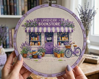 May include: A cross-stitch embroidery hoop featuring a lavender bookstore scene. The design includes a purple storefront with a striped awning, a bicycle, and potted lavender plants. The text "LAVENDER BOOKSTORE" is visible.
