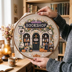 May include: A circular embroidery hoop containing a cross-stitch pattern of a bookshop storefront. The design features the text BOOKSHOP, an OPEN sign, shelves filled with books, pumpkins, and several small white ghosts on a beige fabric.