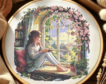 May include: A cross-stitch embroidery hoop with a colorful image of a girl sitting by a window reading a book. The window is surrounded by a lush green vine with pink flowers.