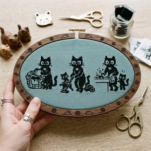 Könnte beinhalten: Ein ovaler hölzerner Stickrahmen mit einem Kreuzstichmuster von schwarzen Katzen in einer häuslichen Umgebung. Die Katzen werden mit verschiedenen Haushaltsgegenständen dargestellt. Der Rahmen wird von einer Hand mit Silberringen gehalten. Weitere Bastelutensilien sind sichtbar.
