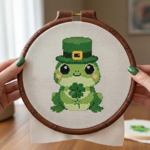 Peut inclure: Broderie au point de croix d'une grenouille verte portant un chapeau de leprechaun et tenant un trèfle à quatre feuilles. La grenouille est centrée dans un cercle à broder en bois. Le fond est en tissu neutre.