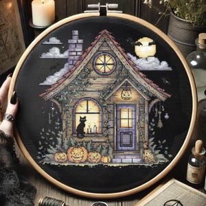 May include: A cross-stitch embroidery of a spooky cottage scene. The design features a wooden house with a pumpkin on the door, a black cat in the window, and pumpkins in front. The background is black with a full moon and clouds.