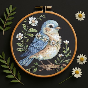 May include: A bluebird with a brown breast sits on a branch surrounded by white daisies. The bird is embroidered on a black fabric and framed in a wooden hoop.