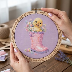May include: Cross-stitch embroidery in a wooden hoop. A yellow chick sits in a pink polka-dot wellington boot filled with flowers. The background is light purple fabric. The wooden hoop has a floral design.