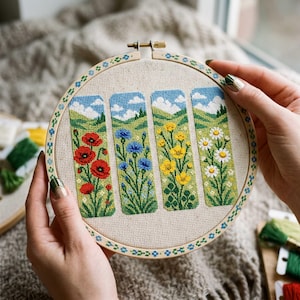 May include: A round wooden embroidery hoop containing a piece of light beige fabric with four vertical cross-stitch panels. Each panel depicts a different wildflower scene with red poppies, blue cornflowers, yellow buttercups, and white daisies.