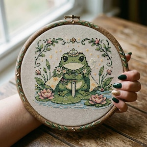 Puede incluir: Bordado de punto de cruz de una rana verde con corona y capa, rodeada de flores y follaje. La obra de arte está enmarcada en un aro de madera redondo con un patrón de hojas decorativas. La rana está sentada en un estanque con nenúfares.