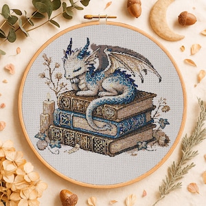Peut inclure: Broderie au point de croix d'un dragon endormi sur une pile de livres. Le dragon est blanc et bleu, avec des ailes détaillées. Les livres sont marron et bleu, dans un cercle à broder en bois.