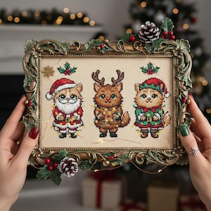 Peut inclure: Broderie au point de croix représentant trois chats costumés pour Noël : le Père Noël, un renne et un elfe. L'œuvre encadrée a un cadre doré orné de houx et de pommes de pin. L'œuvre est tenue par une personne.