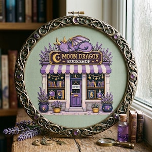 Peut inclure: Une broderie au point de croix représentant une librairie nommée Moon Dragon Bookshop, avec un dragon violet sur le toit, un auvent rayé et de la lavande, encadrée dans un cercle en métal orné de cabochons violets et d'étoiles.