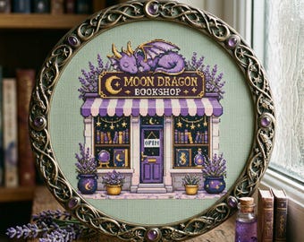 May include: A cross-stitch embroidery of a bookshop named Moon Dragon Bookshop, featuring a purple dragon on the roof, a striped awning, and lavender plants, framed in an ornate circular metal hoop with purple cabochon accents and star motifs.