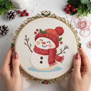 May include: A cross-stitch embroidery of a snowman wearing a red hat and scarf, framed in a gold-colored hoop. The snowman has a carrot nose, twig arms, and a smiling face. The background is white with decorative elements.