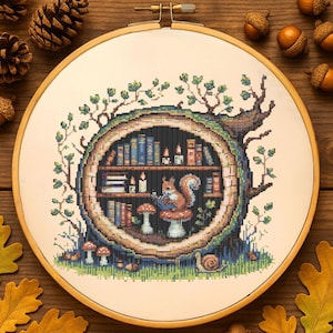 Pode incluir: Bordado em ponto cruz num bastidor de madeira. Um esquilo lê numa biblioteca num tronco de árvore, com livros, velas e cogumelos. O design inclui um caracol e folhas, com uma paleta de cores quentes. O bastidor tem cerca de 20 cm de diâmetro.