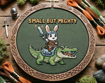 以下が含まれることがあります： 木製のフレームに入ったクロスステッチ刺繍。デザインは、剣を持った緑色のワニに乗るウサギの戦士で、「SMALL BUT MIGHTY」の文字が入っています。背景は濃い緑色の生地です。