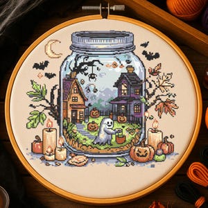 Könnte beinhalten: Kreuzstich-Stickerei mit einer Halloween-Szene in einem Einmachglas. Die Szene zeigt einen Geist, Kürbisse, Häuser und Fledermäuse. Die Stickerei befindet sich in einem Holzrahmen, mit Kerzen und Herbstlaub um das Glas herum.
