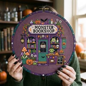 Peut inclure: Un tambour à broder circulaire contenant un motif au point de croix d'une devanture intitulée MONSTER BOOKSHOP. Le design présente un bâtiment violet avec une porte verte, un personnage type Frankenstein, des citrouilles et des livres.