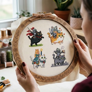 May include: A cross-stitch embroidery hoop with four cat designs dressed as knights and royalty. The cats are riding on other cats. The wooden hoop has a floral and leaf design around the edge and a velvet ribbon. The embroidery is on a cream-colored fabric.