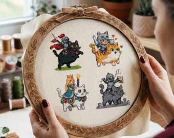 Op de afbeelding: Een borduurring met vier grillige kattenontwerpen. Elke kat is verkleed als ridder, prinses of krijger, rijdend op andere katten. De houten ring heeft een bloemen- en bladerontwerp rond de rand en een fluwelen lint.