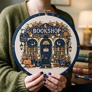 May include: A circular embroidery hoop containing a cross-stitch design of a bookshop storefront. The scene features a dark blue building with the word BOOKSHOP, an OPEN sign, a jack-o'-lantern, stacks of books, and floral accents in orange and green.
