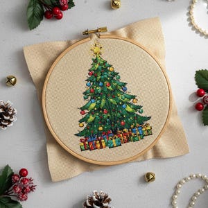 Peut inclure: Une broderie au point de croix d'un sapin de Noël dans un cercle à broder en bois. L'arbre est vert avec des ornements colorés et une étoile dorée. Des cadeaux sont au pied de l'arbre. Le fond est un tissu beige.