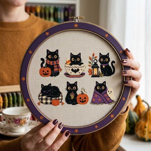 Könnte beinhalten: Ein runder Stickrahmen mit einem Kreuzstichmuster auf beigem Stoff. Das Design zeigt sechs schwarze Katzen in verschiedenen Halloween-Szenen, darunter Kürbisse, eine Teetasse, eine Laterne, ein kariertes Kissen und eine lila Decke mit einem Geist.