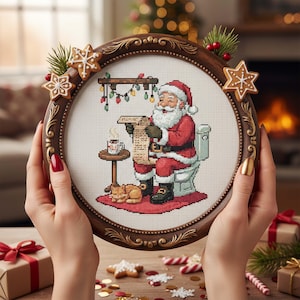May include: A framed cross-stitch depicts Santa Claus on a toilet, reading a list. The round, brown frame is adorned with gingerbread cookies and greenery. A cat sleeps on a red rug. Christmas lights and a mug are also visible. The overall theme is Christmas.