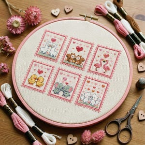 May include: A cross-stitch embroidery hoop featuring six small, framed designs of pairs of animals, including llamas, owls, flamingos, ducks, elephants, and cats, each with a heart. The hoop is pink, and the thread colors are pink, white, and brown.