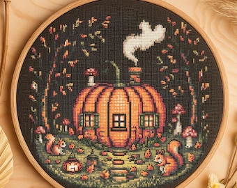May include: Cross-stitch embroidery in a wooden hoop. The design features an orange pumpkin house with a chimney, windows, and a door. Squirrels, mushrooms, and trees surround the house. A white ghost floats above the chimney.