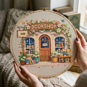 Puede incluir: Un bastidor de bordado de punto de cruz con una representación detallada de una librería. El diseño incluye una puerta de madera, ventanas arqueadas y la palabra "BOOKSHOP" sobre la entrada. La escena está adornada con flores y mariposas, creando una estética encantadora y colorida.