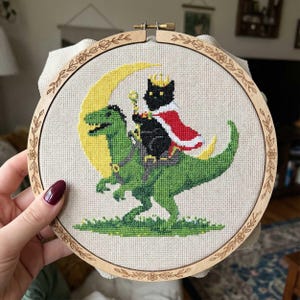 May include: Cross-stitch embroidery in a wooden hoop. The design features a black cat wearing a crown and cape, riding a green dinosaur in front of a yellow crescent moon. The hoop is decorated with floral patterns.