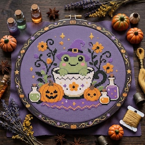 May include: A circular embroidery hoop containing a cross-stitch pattern of a green frog wearing a purple witch hat inside a white teacup. The design includes orange jack-o'-lanterns, small potion bottles, and yellow flowers on a purple fabric.