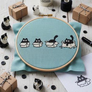 May include: A cross-stitch embroidery hoop featuring black cats in boxes on a light blue fabric. The wooden hoop is surrounded by small black buttons, gift boxes, and ceramic cat figurines. A black marker and spool of thread are also present.