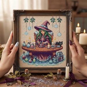 Op de afbeelding: Kruissteekkunst van een heks in een bordeauxrode jurk en hoed, met groen haar, zittend aan een tafel met kaarsen, drankjes en cadeaus. Twee zwarte katten zitten op de tafel. Het kunstwerk zit in een donkerbruine, sierlijke lijst.