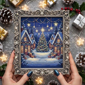 May include: A framed cross-stitch depicting a winter village scene with snow-covered houses, a decorated Christmas tree, and a starry night sky. The ornate silver frame is embellished with crystals. The overall theme is festive and seasonal.