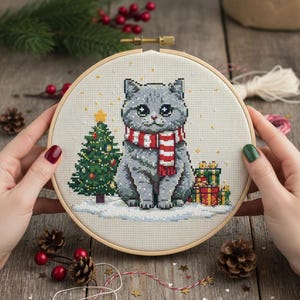 May include: A cross-stitch embroidery hoop showing a grey cat wearing a red and white striped scarf. The cat is in a snowy scene with a Christmas tree and wrapped gifts. The embroidery is set in a wooden hoop.
