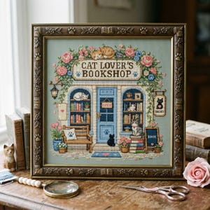 May include: A framed cross-stitch embroidery depicting a storefront titled Cat Lover's Bookshop. The scene features a blue door, bookshelves filled with books, various cats, and floral accents in pink, blue, and green, all encased in a brown frame.