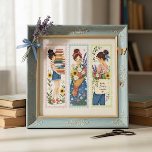 Pode incluir: Uma obra de arte em ponto cruz emoldurada com três painéis de mulheres com livros e flores. A moldura azul clara tem bordas decorativas. Um maço de lavanda e uma fita azul estão presos. Borboletas também estão presas à moldura.