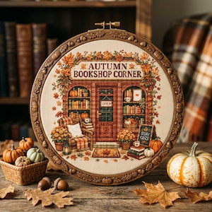 May include: A circular embroidery hoop frame containing a cross-stitch design of a storefront labeled "AUTUMN BOOKSHOP CORNER." The scene features bookshelves, a cozy chair, pumpkins, and fall foliage in shades of orange, brown, and green.