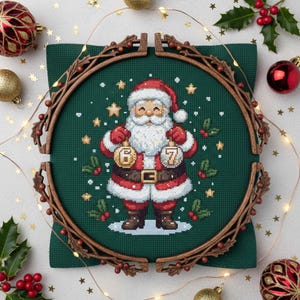 Könnte beinhalten: Ein Kreuzstich von Weihnachtsmann, der Ornamente mit den Zahlen 6 und 7 hält. Er trägt einen roten Anzug und Hut mit weißem Bart auf grünem Hintergrund. Der Kreuzstich ist in einem hölzernen Stickrahmen gerahmt, mit Weihnachtsdekorationen und Lichtern.