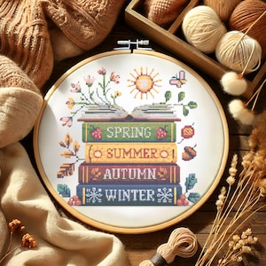 May include: A cross-stitch embroidery hoop featuring a design of stacked books representing the seasons: Spring, Summer, Autumn, and Winter. The design includes a sun, flowers, and a butterfly, all in vibrant colors.