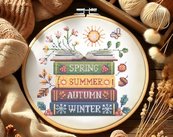 May include: A cross-stitch embroidery hoop featuring a design of stacked books representing the seasons: Spring, Summer, Autumn, and Winter. The design includes a sun, flowers, and a butterfly, all in vibrant colors.
