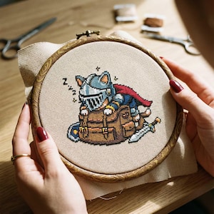 May include: Cross-stitch embroidery of a sleeping cat knight in a helmet and cape, on a brown backpack. The design is in a wooden embroidery hoop, held by hands with red nail polish. Scissors and thread spools are in the background.