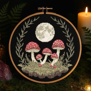 May include: A black embroidery hoop with a cross-stitch design of a full moon and red mushrooms in a forest setting.