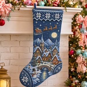 Puede incluir: Un calcetín navideño azul oscuro con un diseño detallado de punto de cruz de un pueblo nevado de noche. La escena incluye una luna llena, un puente sobre un arroyo, un muñeco de nieve y a Santa Claus en un trineo con renos sobre las casas.