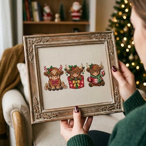 May include: A framed cross-stitch embroidery on beige fabric featuring three brown Highland cows with Christmas decorations. The first cow wears a red bow, the second sits in a red gift box, and the third wears a green scarf and holds a stocking.