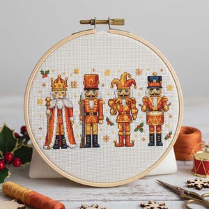 Könnte beinhalten: Ein Kreuzstich-Stickrahmen mit vier Spielzeugsoldaten-Nussknackern in Orange, Gelb und Schwarz. Jeder Nussknacker hat eine einzigartige Uniform und einen Hut. Der Hintergrund ist weiß, mit Schneeflocken und Stechpalmenzweigen.