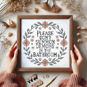 May include: A cross-stitch embroidery with a floral wreath and the text "Please don't summon demons in the bathroom".