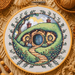 Può includere: Ricamo a punto croce in un telaio di legno. Il disegno raffigura una casa di hobbit con una porta blu, due finestre rotonde e un sentiero. La scena è ambientata in verdi colline, con un albero e un cielo nuvoloso. Diametro: circa 20 cm.
