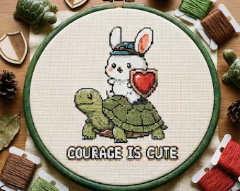 以下が含まれることがあります： 木製のフレームに入ったクロスステッチ刺繍。カメに乗った漫画のウサギがハート型の盾を持っています。「COURAGE IS CUTE」の文字が刺繍されています。デザインは、緑と茶色の刺繍糸、木製の盾、装飾的な要素で囲まれています。